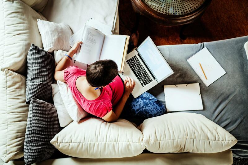 Aerial view of a mother sat on her couch, looking through a booklet and using her laptop while working from home.