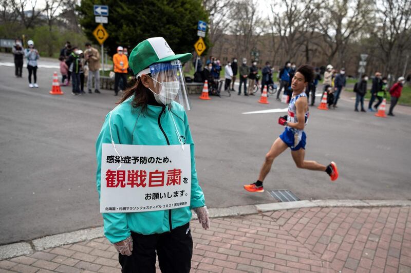 A volunteer (L) holds a placard asking people to refrain from watching the competition to prevent the spread of the Covid-19 coronavirus while an athlete (R) competes in the half-marathon race which doubles as a test event for the 2020 Tokyo Olympics, in Sapporo on May 5, 2021. / AFP / POOL / Charly TRIBALLEAU
