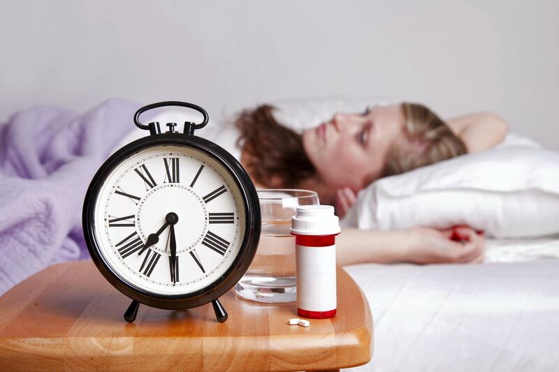 Woman lays sleepless in bed (blurred) with an alarm clock, pills, pill bottle, and water on the nightstand; copy space 