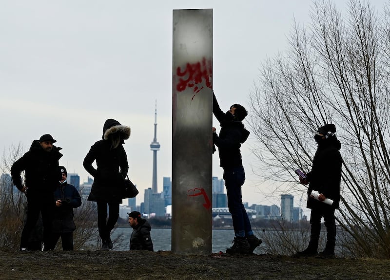 A man tries to clean graffiti from the vandalized monolith as people look at a metallic monolith that appeared on the shorelines of Humber Bay Park during the COVID-19 pandemic in Toronto on Friday, Jan. 1, 2021. (Nathan Denette/The Canadian Press via AP)