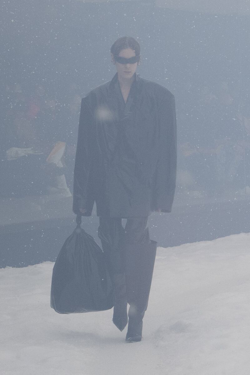 A model in an oversized jacket and boots clutches a black plastic bag.