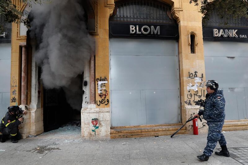 FILE - In this Tuesday, Feb. 11, 2020 file photo, a firefighter looks before entering a bank that was set on fire by anti-government protesters, as a riot policeman passes by, during a protest in downtown Beirut, Lebanon. Fitch Ratings said Tuesday, Feb. 18, 2020, that Lebanon's financial position points to a likely restructuring of the country's massive debt and financial sector as the Arab nation passes though its worst financial and economic crisis in decades. (AP Photo/Hussein Malla, File)