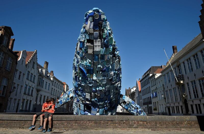 Children sit near a 12-metre installation depicting a whale, made up of five tons of plastic waste pulled out of the Pacific Ocean, is displayed in Brugges, on July 14, 2018 for the 2018 Bruges Triennial.  / AFP / JOHN THYS
