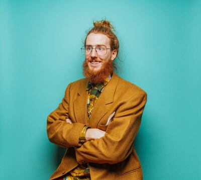 Studio. photograph of Hipster man on blue background. With beard and hair in a bun. Wearing a vintage blazer and multi-coloured shirt, and eyeglasses. Arms crossed. Getty Images