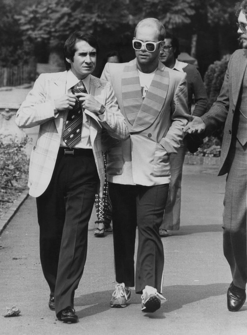 Elton John, in a jacket with a striped lapel and white sunglasses, with his manager John Reid on their way to a luncheon in honour of Buddy Holly in Holland Park, London on September 8, 1976. Getty Images