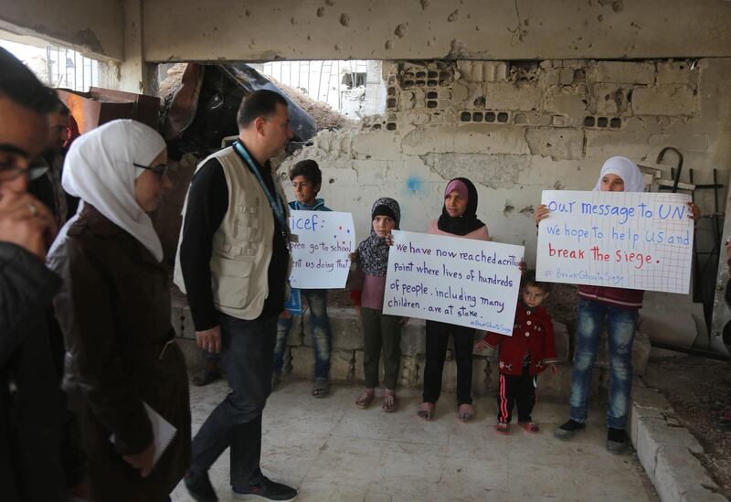 Syrian children hold placards as members of an aid convoy enter a building during a humanitarian delivery in the al-Nashabia town, in the rebel-held Eastern Ghouta region, east of the capital Damascus on November 28, 2017.
The convoy carrying food and medical aid entered Syria's rebel-held Eastern Ghouta in a rare humanitarian delivery that comes after days of heavy bombardment. / AFP PHOTO / Amer ALMOHIBANY