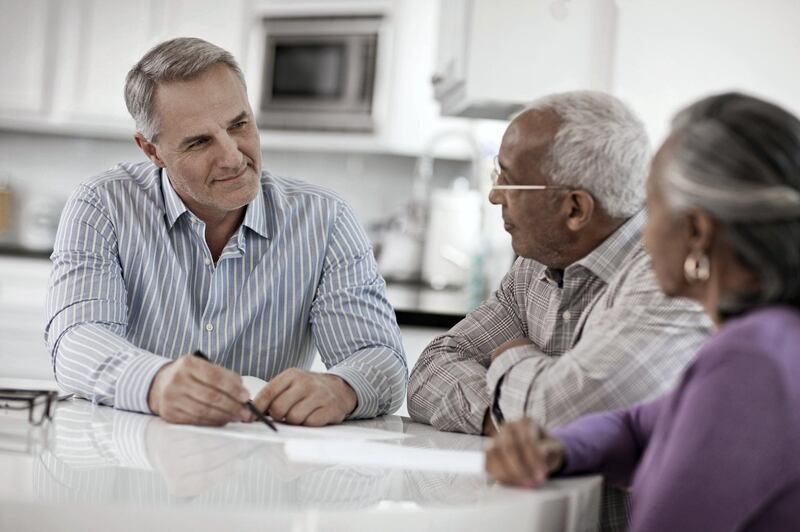 Three people at a table, a senior couple seated with a man using paper and pen to give them information.