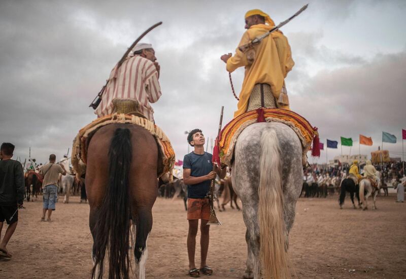 In this Thursday, July 25, 2019 photo, a boy hands a rifle to horse riders waiting for their turn to take part in an equestrian show known as Fantasia or Tabourida, in the coastal town of El Jadida, Morocco.
