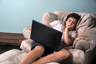 A boy reclining in a chair at home using a laptop computer.  He looks tired and worn out.  A concept on too much screen time and binge watching for children.