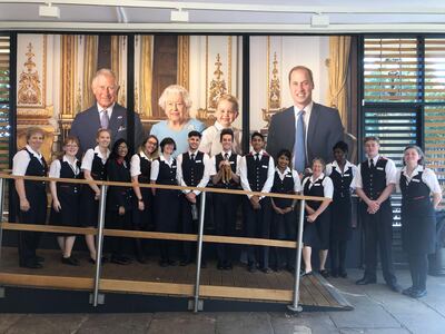 Harriet smiles with palace staff under a photograph of the Queen, Prince Charles, Prince William and Prince George, the British line of succession. Courtesy Royal Collection Trust / © Her Majesty Queen Elizabeth II 2019.