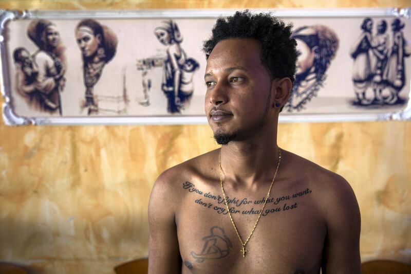 Danny Avram,27,an Eritrean asylum seeker at the Harhara cafe in south Tel Aviv poses with his shirt off to show his tattoo he got in Israel that reads, 'If you don't fight for what you want, don't cry for what your lost " on March 19,2018.He recounted how Beduin smugglers shot him in the leg three times when he was unable to meet their demand for forty thousand dollars. His first year in Israel he could not work because of the injuries, but then he landed a job as a dishwasher. "Why to Rwanda?"he asked. "They can't even take care of their own people there, how are they going to take care of refugees?"(Photo by Heidi Levine for The National).