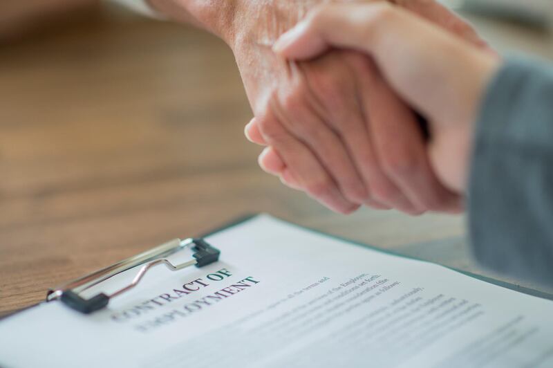Two people are standing at a table, across from each other. Their hands grasp each others in a firm handshake. On the table sits a contract of employment. Getty Images