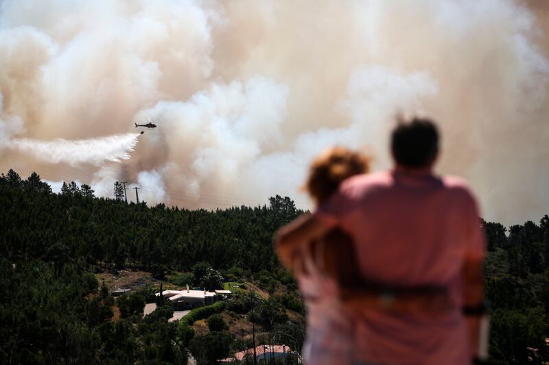 TOPSHOT - A couple watches an helicopter dropping water on a wildfire near Monchique, in Algarve, on August 8, 2018. - Spain and Portugal approached record temperatures at the weekend, with the mercury hitting 46.6 degrees Celsius (116 Fahrenheit) at El Granado in Spain and 46.4 C in Alvega, Portugal, according to the World Meteorological Organisation (WMO). While the deadly hot spell is expected to ease in parts of western Europe in the coming days, firefighters in Spain and Portugal struggled to contain wildfires that have swept southern areas. (Photo by CARLOS COSTA / AFP)