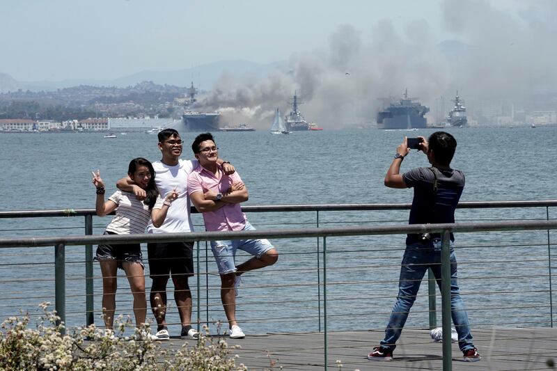 People pose for a picture in front of the San Diego skyline as smoke rises from a fire on board 'USS Bonhomme Richard' at the naval base in San Diego, as seen from Coronado, California, US. Reuters