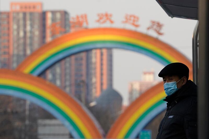 A resident wearing a mask to protect from the coronavirus waits at a bus stop near rainbow decorations on the streets of Beijing on Sunday, Dec. 27, 2020. Beijing has urged residents not to leave the city during the Lunar New Year holiday in February, implementing new restrictions and mass testings after several coronavirus infections last week. (AP Photo/Ng Han Guan)