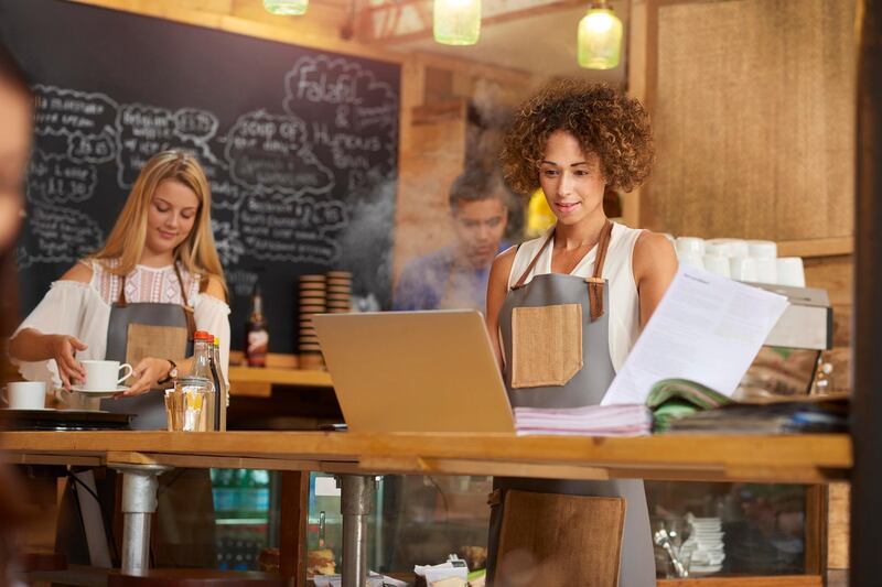 a coffee shop owner checks the latest bills in her bookkeeping folder on the counter of her busy coffee shop. In the background two staff attend to the orders.