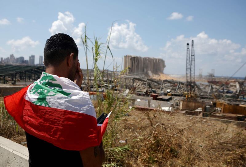 TOPSHOT - A man draped in a Lebanese flag reacts as he stands before the ravaged port of Lebanon's capital Beirut on August 9, 2020, in the aftermath of a colossal explosion that occurred days prior due to a huge pile of ammonium nitrate that had languished for years at a port warehouse. The huge chemical explosion that hit Beirut's port, devastating large parts of the Lebanese capital and claiming over 150 lives, left a 43-metre (141 foot) deep crater, a security official said. The blast Tuesday, which was felt across the country and as far as the island of Cyprus, was recorded by the sensors of the American Institute of Geophysics (USGS) as having the power of a magnitude 3.3 earthquake. / AFP / PATRICK BAZ
