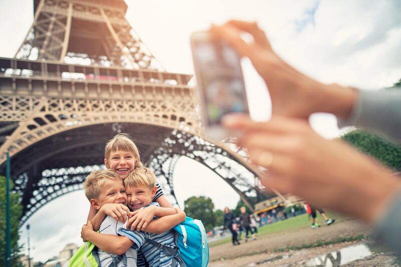 Mother taking picture of  little girl with her younger brothers hugging near The Eiffel Tower. The tower is visible in the background. The kids aged 9 and 6 are hugging and smiling at the camera. Paris, France. Getty Images
