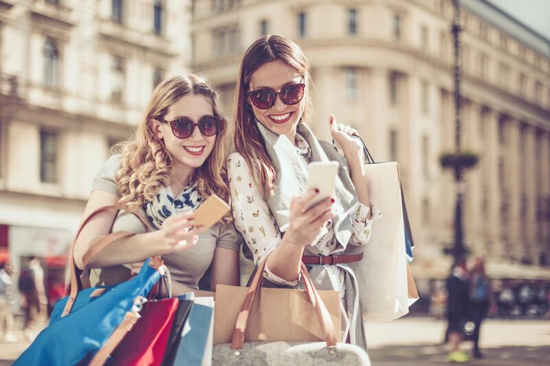 Cheerful beautiful women are shopping in the city. They are using phone and drinking a coffee to go. Getty Images