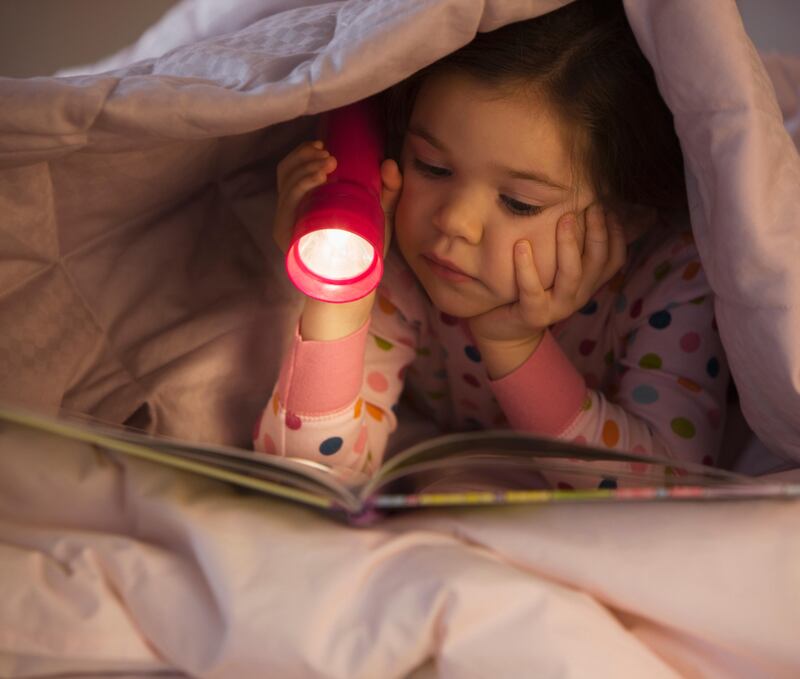Mixed race girl reading book in bed with flashlight. Getty Images