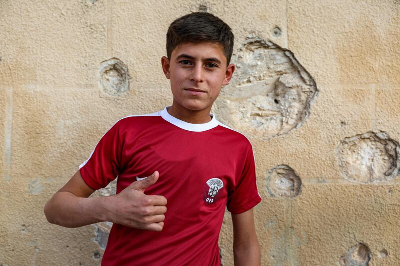 A boy wearing a Qatar jersey poses for a photo next to a wall riddled with bullet holes