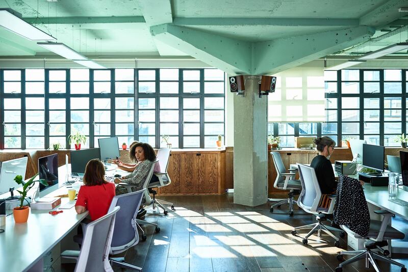 Two female co-workers chatting, mature woman working on computer, sunlight through large window, warehouse style open plan office. Getty Images