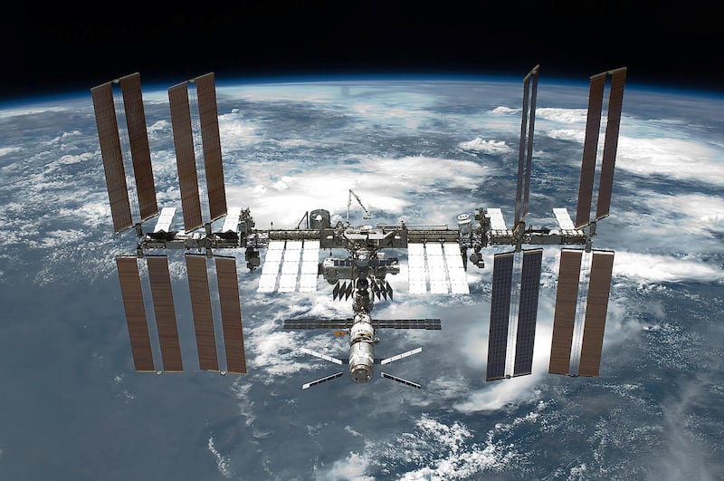 A view of the International Space Station backdropped by Earth. The station has four large, maroon-coloured solar array wings, two on either side of the station, mounted to a central truss structure. Further along the truss are six large, white radiators, three next to each pair of arrays. In between the solar arrays and radiators is a cluster of pressurised modules arranged in an elongated T shape, also attached to the truss. A set of blue solar arrays are mounted to the module at the aft end of the cluster. Wikicommons