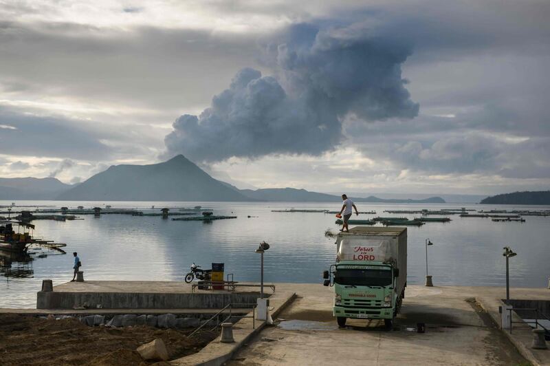 A man clears ash from the roof of his truck, before a plume of steam rising from the Taal volcano, at a fishing harbour in Laurel on January 17, 2020. / AFP / Ed JONES
