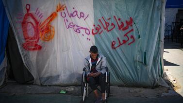A Palestinian youth checks his phone as he sits in a wheelchair by a makeshift tent bearing the Arabic inscription 'Ramadan brings us together - we want Qatayef and biscuits', traditional food made during the Muslim holy fasting month of Ramadan, in Rafah in the southern Gaza Strip on March 12, 2024, amid ongoing battles between Israel and the militant group Hamas.  (Photo by AFP)