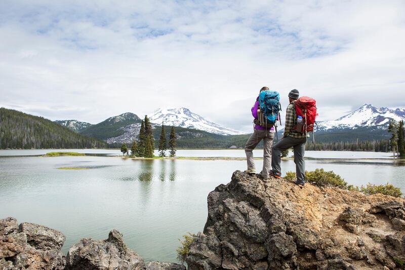 Two Hikers standing on a viewpoint point overlooking a lake with two snowy mountains in the distance. Getty Images