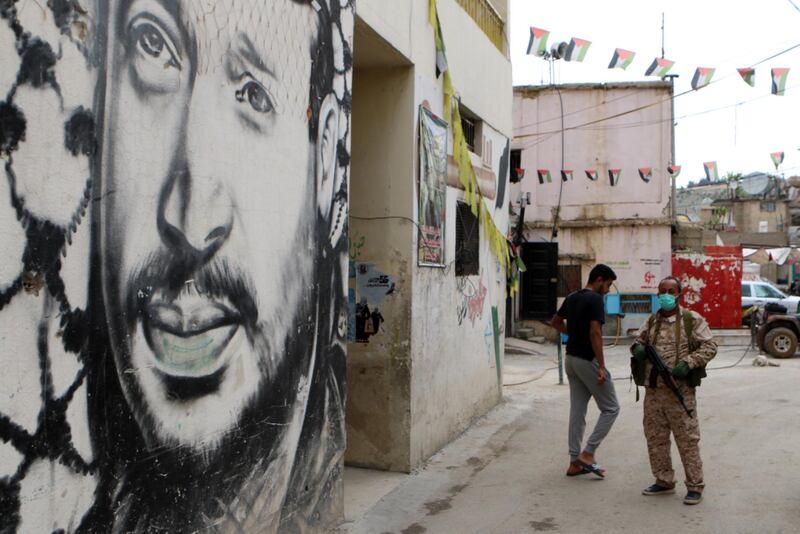 Armed Palestinians wearing protective face masks stand in front of a portrait of their late leader Yasser Arafat at the Wavel Palestinian refugee camp (also known as the Jalil camp) in Lebanon's eastern Bekaa Valley, on April 24, 2020, after cases of infection by the novel coronavirus were detected there.  The residents of the Wavel camp were tested after a member of a household, a Palestinian refugee from Syria, was admitted to the state-run Rafic Hariri hospital in the capital Beirut  for demonstrating COVID-19 symptoms. / AFP / -
