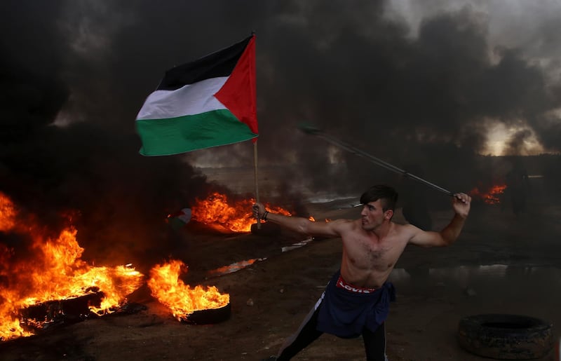 FILE-- In this Friday, Oct. 26, 2018 file photo, a protester hurls stones while holding the Palestinian flag amid burning tires near the fence of the Gaza Strip border with Israel during a protest. Palestinians in the Gaza Strip have coped with shortages of just about everything in more than a decade of border closures -- from chocolate to medicines to fuel and building supplies. Now, the past six months of protests against an Israeli-Egyptian blockade have added an unexpected item to the list of those in short supply: car tires. (AP Photo/Adel Hana, File)