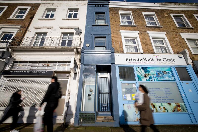 People walk past the front of what is dubbed 'London's thinnest house' (painted blue) in west London on February 5, 2021. Wedged neatly between a doctor's surgery and a shuttered hairdressing salon, the five floor house in Shepherds Bush is just 5ft 6ins (1.6 metres) at its narrowest point and is currently on the market for £950,000 ($1,300,000, 1,100,000 euros). The unusual property, originally a Victorian hat shop with storage for merchandise and living quarters on its upper floors, was built sometime in the late 19th or early 20th century.
 / AFP / Tolga Akmen
