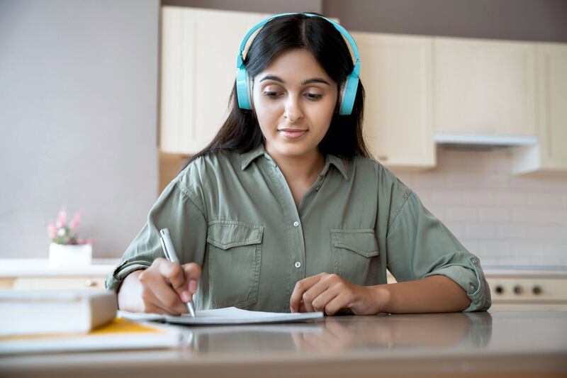 2C9X5GN Indian girl wear headphones writing in notebook listening audio lesson at home.