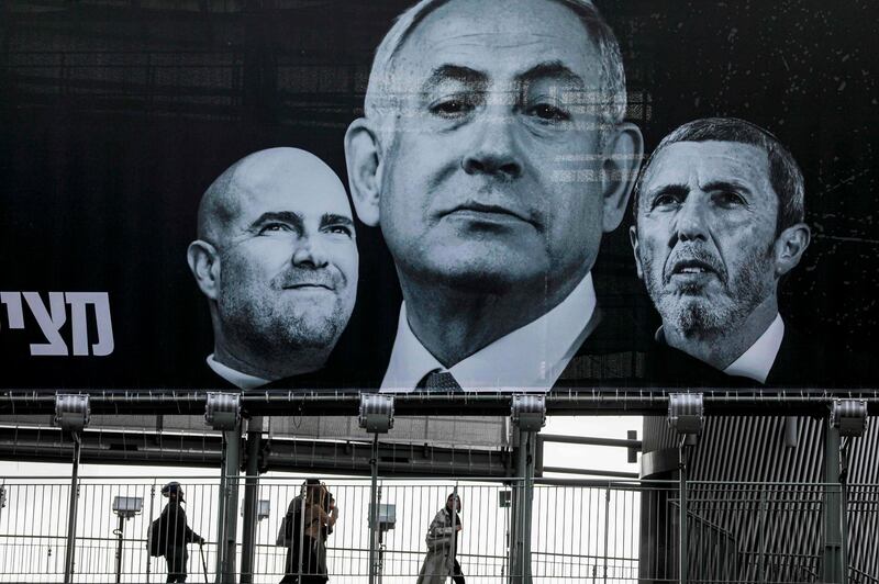walk along a pedestrian overpass beneath a giant Blue and White (Kahol Lavan) alliance electoral billboard showing the face of Israeli Prime Minister Benjamin Netanyahu (C) flanked by Justice Minister Amir Ohana (L) and Education Minister Rafi Peretz (R) in the central Israeli city of Ramat Gan on March 1, 2020, a day before the country's third election in a year.  / AFP / MENAHEM KAHANA
