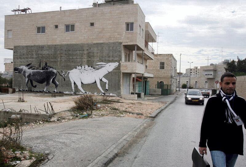 epa01191069 Illusive British graffiti artist named Banksy has painted new works in the West Bank town of Bethlehem, which might include a huge painting of two donkeys with their tails entwined pulling each other, as seen on the side of a house in Bethlehem as a Palestinian walks past on 04 December 2007. The white donkey has what appears to be a pastoral palestinian village on its back and the black donket carries on its back what appears to be a modern Israeli town or cityscape. Banksy's works are not signed.  EPA/JIM HOLLANDER *** Local Caption *** 01191069