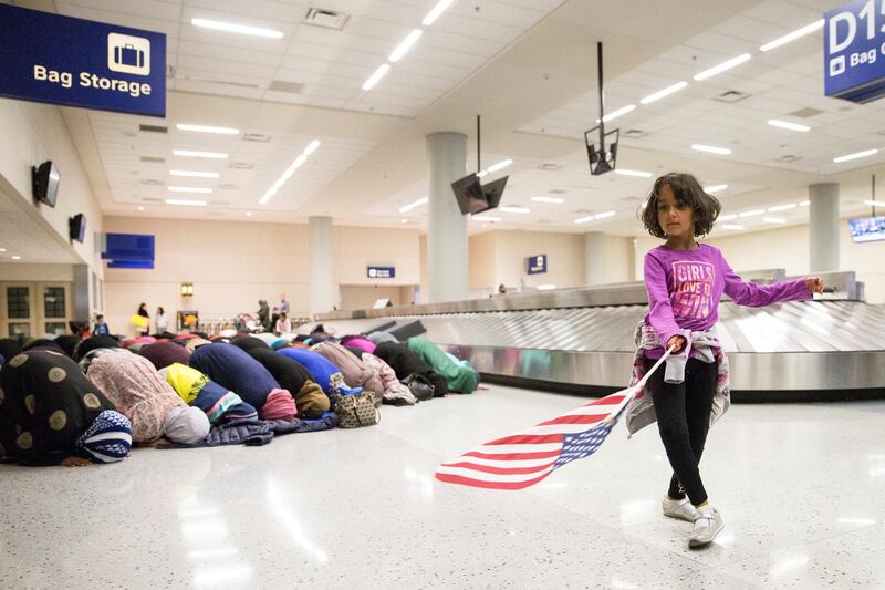 A young girl dances with an American flag in baggage claim while women pray behind her during a protest against the travel ban imposed by US President Donald Trump's executive order, at Dallas/Fort Worth International Airport in Dallas, Texas, on January 29, 2017. Reuters