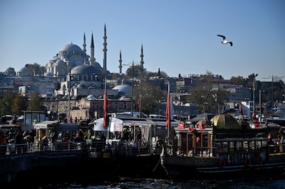 People walk by fish and bread boats, specialised in fish sandwiches, near the Galata bridge with the Suleymaniye mosque in the background, on December 6, 2019, during a sunny day at the Eminonu district in Istanbul. / AFP / Ozan KOSE
