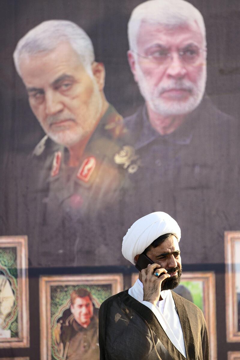 A Muslim cleric speaks on the phone in front of a poster depicting slain Iranian general Qasem Soleimani (L) and slain Iraqi commander Abu Mahdi al-Muhandis during a military parade for Hashed al-Shaabi (Popular Mobilisation) paramilitary force in the southern Iraqi city of Basra on June 14, 2020, marking the sixth anniversary of its founding after Iraq's top Shiite cleric Grand Ayatollah Ali Sistani called to defend the country from the Islamic State group (IS). (Photo by Hussein FALEH / AFP)