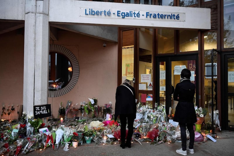 People look at flowers at the entrance of a middle school in Conflans-Sainte-Honorine, 30kms northwest of Paris, on October 17, 2020, after a teacher was decapitated by an attacker who has been shot dead by policemen.  The man suspected of beheading on October 16 ,2020 a French teacher who had shown his students cartoons of the prophet Mohammed was an 18-year-old born in Moscow and originating from Russia's southern region of Chechnya, a judicial source said on October 17. Five more people have been detained over the murder on October 16 ,2020 outside Paris, including the parents of a child at the school where the teacher was working, bringing to nine the total number currently under arrest, said the source, who asked not to be named. The attack happened at around 5 pm (1500 GMT) near a school in Conflans-Sainte-Honorine, a western suburb of the French capital. The man who was decapitated was a history teacher who had recently shown caricatures of the Prophet Mohammed in class. / AFP / Bertrand GUAY
