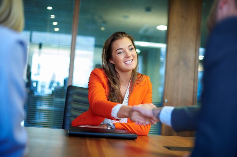 a young graduate sits across the table from her interview panel  full of confidence and positivity energy . She is holding her cv and smiling at the interview panel before her and shakes hands with the new boss.She is wearing blue trousers with an orange suit jacket . in the foreground we can see the back of two of the panel , a man and a woman.