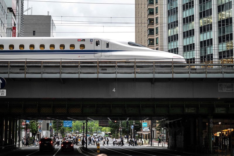 People cross a street while a shinkansen, or high speed bullet train, leaves Tokyo on June 16, 2021. / AFP / Charly TRIBALLEAU
