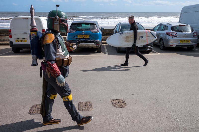 A man dressed as Boba Fett from Star Wars walks past a surfer on the seafront