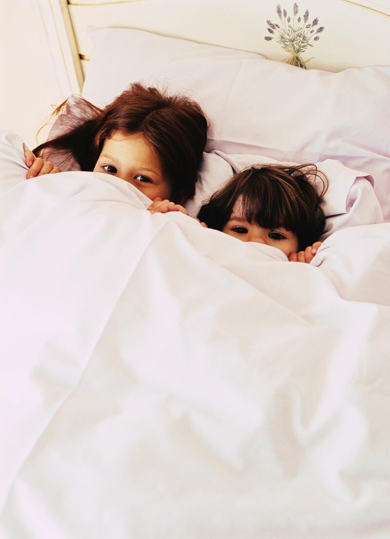 Portrait of two children (4-10) lying in bed holding a cover over their faces. Getty Images