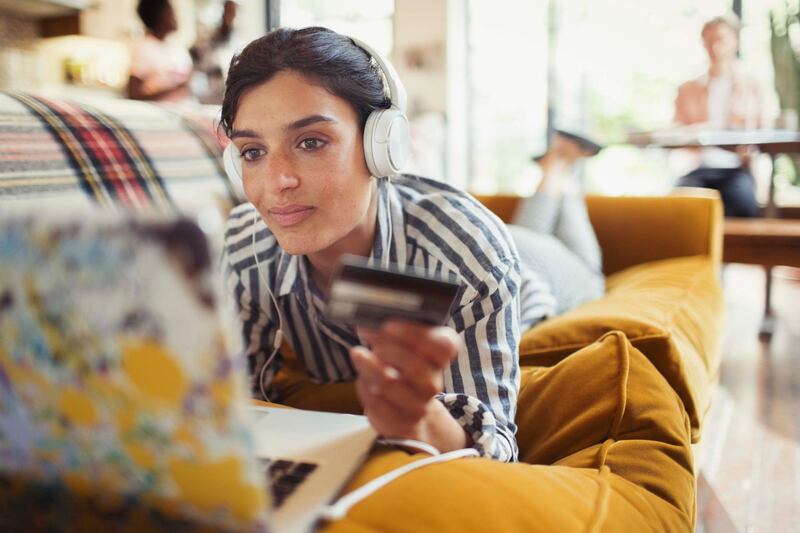 Young woman with credit card and headphones online shopping at laptop on living room sofa. Getty Images