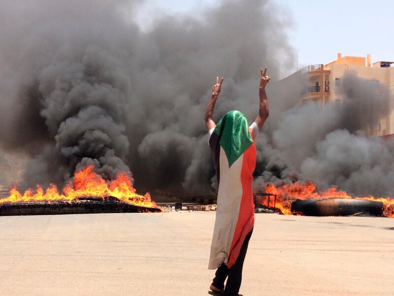 FILE - In this June 3, 2019 file photo, a protester wearing a Sudanese flag flashes the victory sign in front of burning tires and debris on road 60, near Khartoum's army headquarters, in Khartoum, Sudan,  Sudanese prosecutors say the country's ruling generals did not order the deadly break-up of a protest camp last month. Prosecutor Fathel-Rahman Said announced that eight officers from the paramilitary Rapid Support Forces had exceeded their orders when they led RSF troops to clear a pro-democracy sit-in on June 3 in the capital, Khartoum. He said security forces were told to clear a lawless area close to the sit-in, not to raze the protest camp. Said, who heads a committee investigating the dispersal, said on Saturday, July 27,  that the officers, including a major general, have been accused of crimes against humanity. (AP Photo)