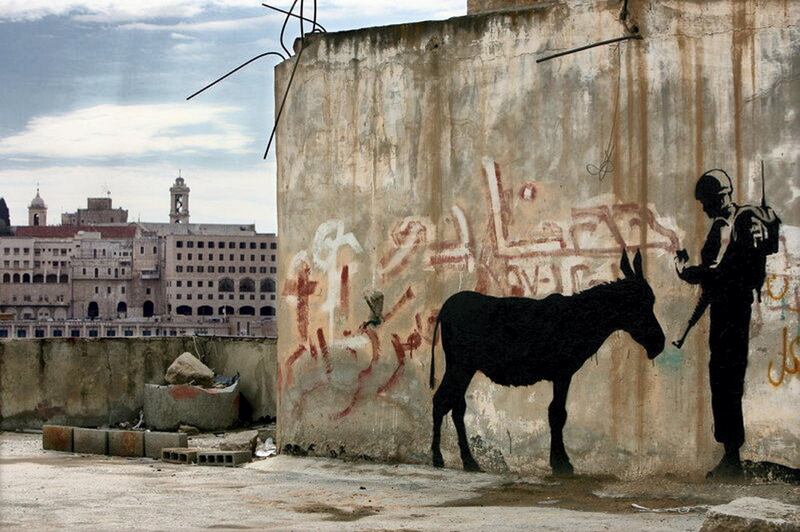 epa01191023 Illusive British graffiti artist named Banksy has painted new works in the West Bank town of Bethlehem including  this stenciled work showing an Israeli soldier asking a donkey for it's identity card, on 04 December 2006. Behind, at left, is the old town of Bethlehem, with towers from the Church of the Nativity, the traditional birthplace of Jesus Christ. These new works are being dubbed 'West Banksy,' and some are painted on the controversial Israeli 'separation barrier,' or wall, that surrounds Bethlehem and cuts its residents off from nearby Jerusalem.  EPA/JIM HOLLANDER *** Local Caption *** 01191023