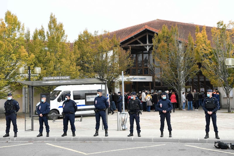 French CRS police officers stand as adults and children gather in front of flowers displayed at the entrance of a middle school in Conflans-Sainte-Honorine, 30kms northwest of Paris, on October 17, 2020, after a teacher was decapitated by an attacker who has been shot dead by policemen.  The man suspected of beheading on October 16 ,2020 a French teacher who had shown his students cartoons of the prophet Mohammed was an 18-year-old born in Moscow and originating from Russia's southern region of Chechnya, a judicial source said on October 17. Five more people have been detained over the murder on October 16 ,2020 outside Paris, including the parents of a child at the school where the teacher was working, bringing to nine the total number currently under arrest, said the source, who asked not to be named. The attack happened at around 5 pm (1500 GMT) near a school in Conflans Saint-Honorine, a western suburb of the French capital. The man who was decapitated was a history teacher who had recently shown caricatures of the Prophet Mohammed in class. / AFP / Bertrand GUAY
