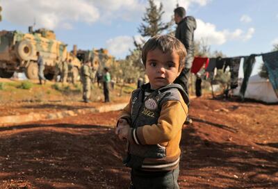 A displaced Syrian child looks to the camera as Turkish soldiers gather near the town of Batabu on the highway linking Idlib to the Syrian Bab al-Hawa border crossing with Turkey, on March 2, 2020. The Syrian government pledged to repel Turkish forces attacking its Russia-backed troops in northwestern Syria as tensions spike between Damascus and Ankara. Since December, Syrian regime forces have led a deadly military offensive against the last major opposition stronghold of Idlib, where Turkey supports some rebel groups. Damascus said it shot down three Turkish drones in the region the day before, while two Syrian warplanes were downed though the pilots escaped unharmed. / AFP / AAREF WATAD
