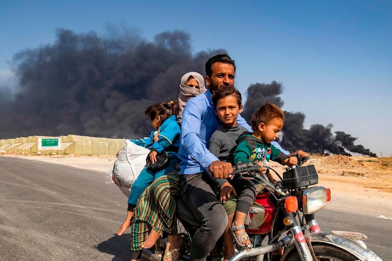 TOPSHOT - Displaced people, fleeing from the countryside of the Syrian Kurdish town of Ras al-Ain along the border with Turkey, ride a motorcycle together along a road on the outskirts of the nearby town of Tal Tamr on October 16, 2019 as they flee from the Turkish "Peace Spring" military operation, with smoke plumes of tire fires billowing in the background to decrease visibility for Turkish warplanes in the area.  / AFP / Delil SOULEIMAN
